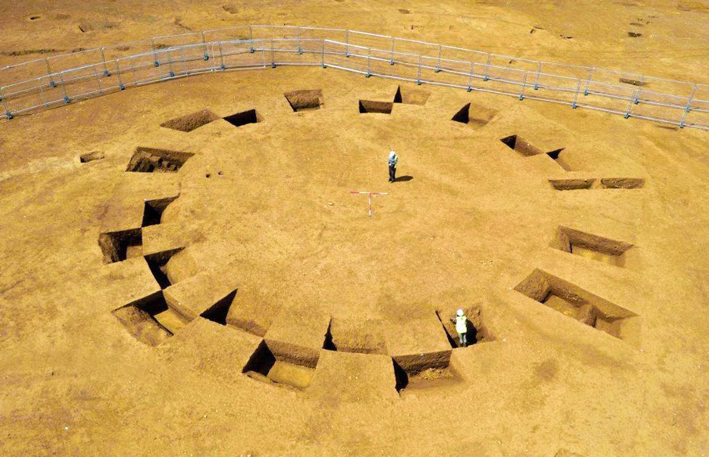 Rectangular holes dug in the ground in the shape of a circle one person standing in the centre of the circle one person standing in one of the holes