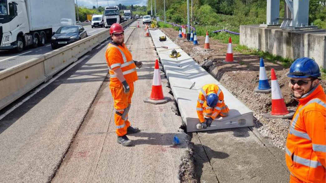 Workers wearing high vis and hardhats on the side of the M6 smiling at the camera.